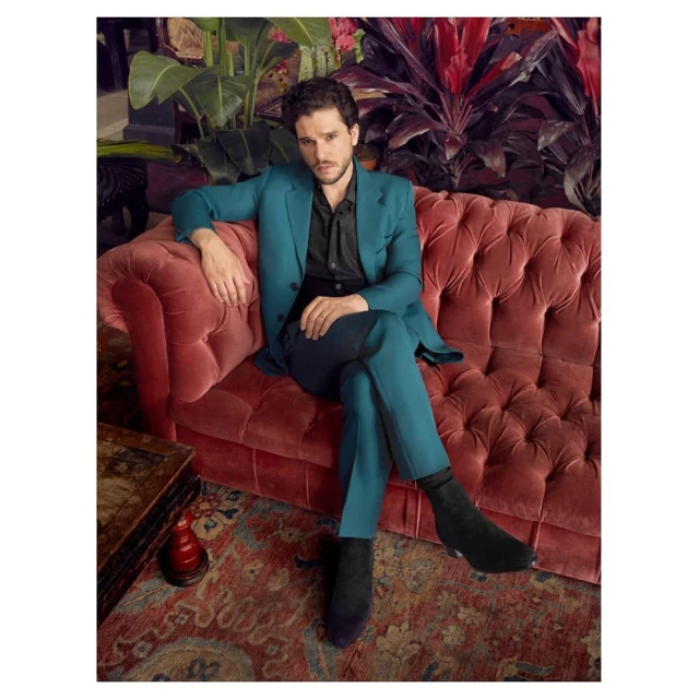Adult man seated on an upholstered sofa, wearing a teal suit and black boots, in an interior with a patterned rug and houseplants.