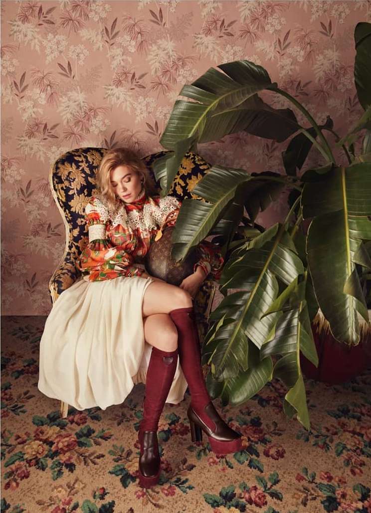 Fashionable woman sits in an ornate chair, holding a bottle, in a room with floral wallpaper and a lush potted plant.
