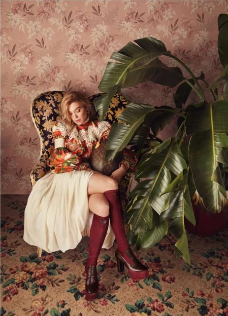 Fashionable woman sits in an ornate chair, holding a bottle, in a room with floral wallpaper and a lush potted plant.