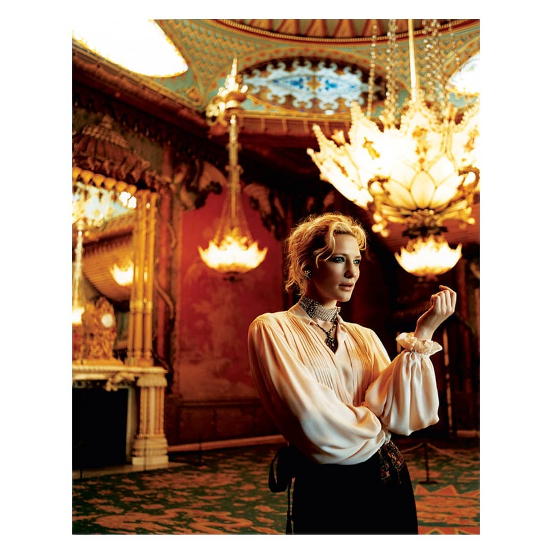 Indoor portrait of an adult female in a vintage blouse standing in an ornate room with chandeliers, decorative walls, and patterned carpet.