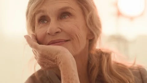 Close-up portrait of a smiling elderly woman with wavy blonde hair resting her face on her hand, set against a blurred background.