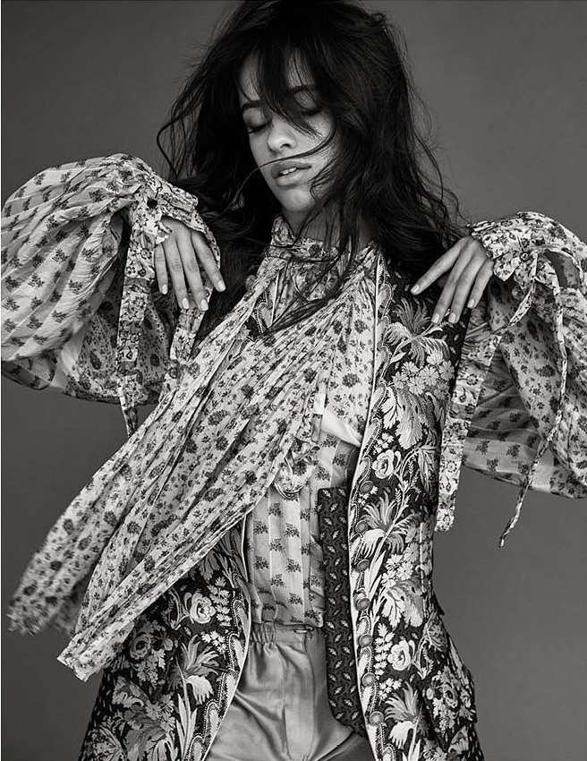 Black and white portrait of a woman with dark hair wearing a patterned jacket and blouse, hands raised, and eyes closed in a studio setting.