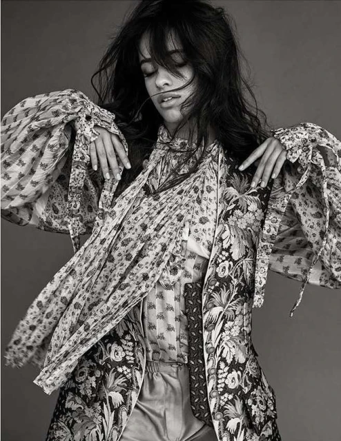 Black and white portrait of a woman with dark hair wearing a patterned jacket and blouse, hands raised, and eyes closed in a studio setting.