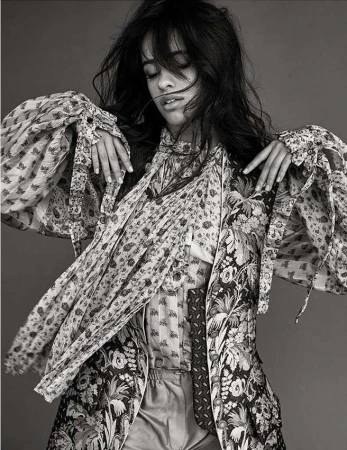 Black and white portrait of a woman with dark hair wearing a patterned jacket and blouse, hands raised, and eyes closed in a studio setting.