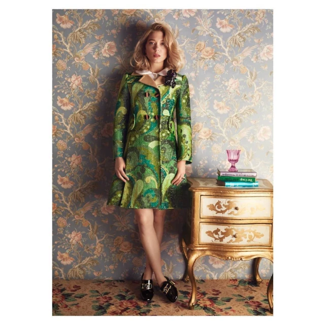Woman in green patterned coat standing in front of floral wallpaper, beside a small gold table with books and a pink glass.