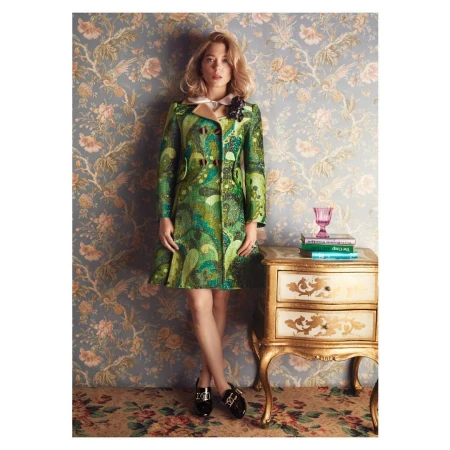Woman in green patterned coat standing in front of floral wallpaper, beside a small gold table with books and a pink glass.