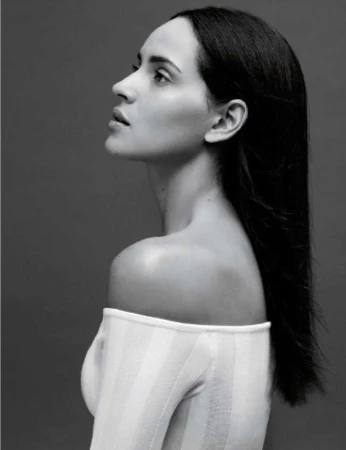 Profile portrait of a woman with long dark hair wearing a white off-the-shoulder top against a grey background