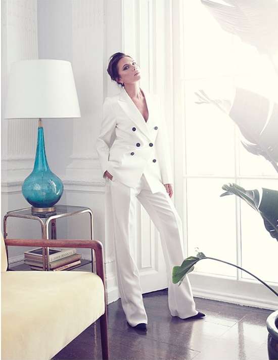 A woman in a white pantsuit poses in an airy, bright room near a window with a lamp on a glass table and green plant.