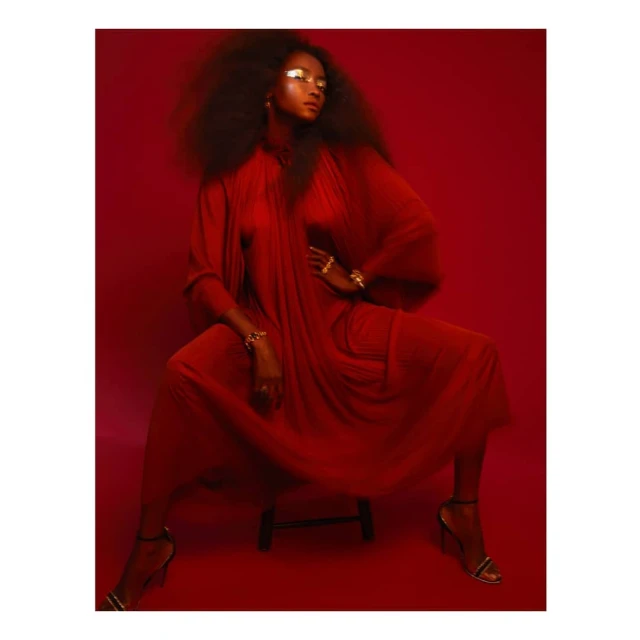 Woman in flowing red gown with dramatic eye makeup sitting on a stool against a red backdrop in a studio portrait.