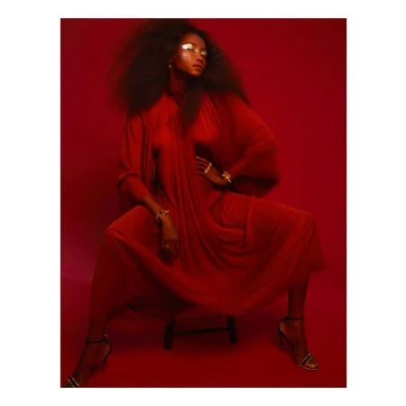 Woman in flowing red gown with dramatic eye makeup sitting on a stool against a red backdrop in a studio portrait.