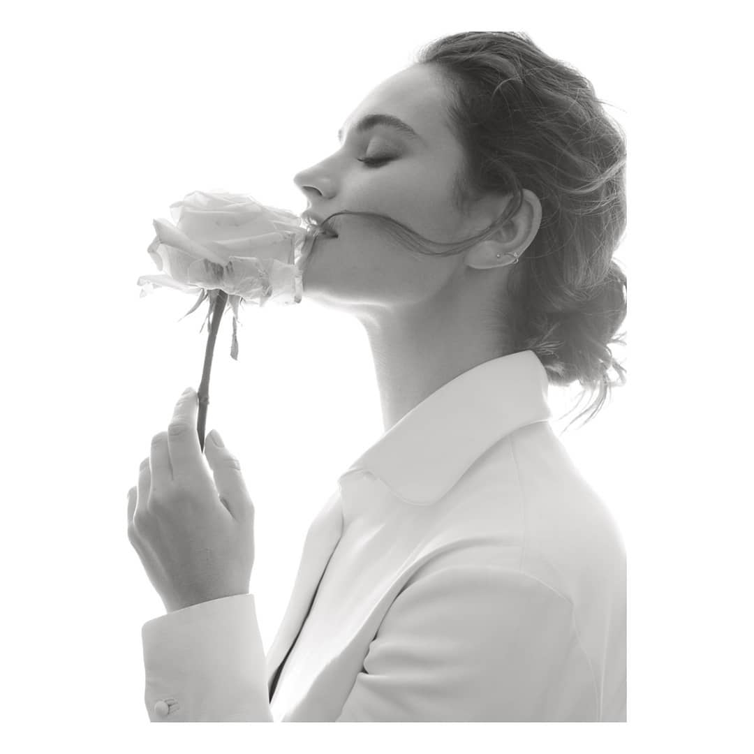 Monochromatic portrait of a woman in a white suit smelling a pale rose, eyes closed, in a close-up shot, and high-key lighting.