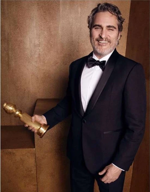 Actor Joaquin Phoenix in tuxedo holding a golden statuette award against a brown textured background, smiling with salt and pepper hair.