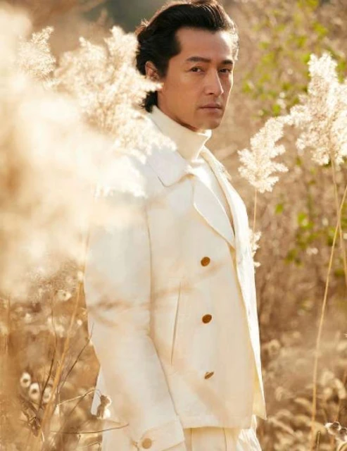 Elegant Asian man in a white double-breasted coat poses in a grassy field, showcasing fashion and outdoor style with a serene expression.