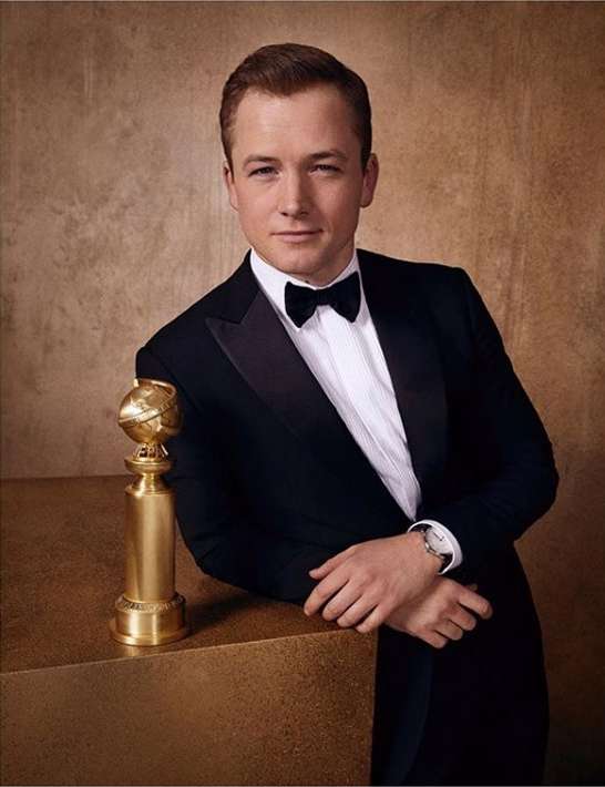 Taron Egerton poses in a tuxedo with a Golden Globe award, set against a textured gold backdrop, capturing a moment of achievement.