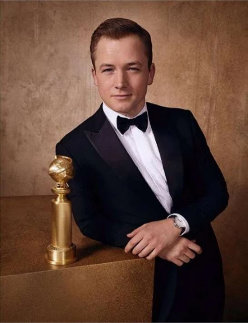 Taron Egerton poses in a tuxedo with a Golden Globe award, set against a textured gold backdrop, capturing a moment of achievement.