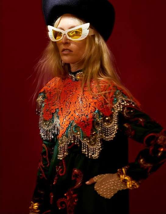 Woman in ornate vintage couture, wearing beret and winged glasses, poses in front of a red backdrop, showcasing intricate details.