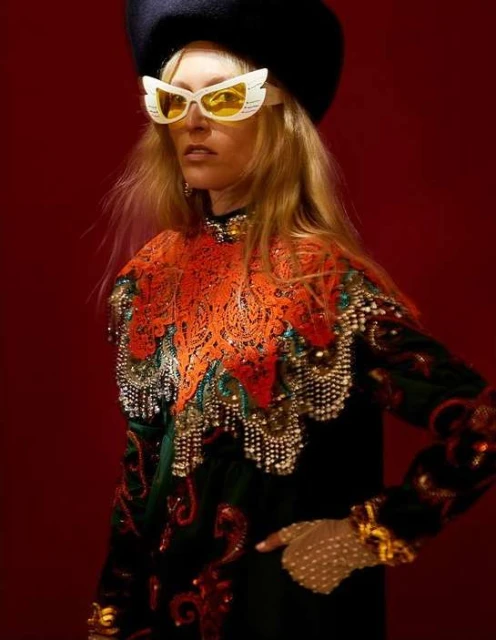 Woman in ornate vintage couture, wearing beret and winged glasses, poses in front of a red backdrop, showcasing intricate details.
