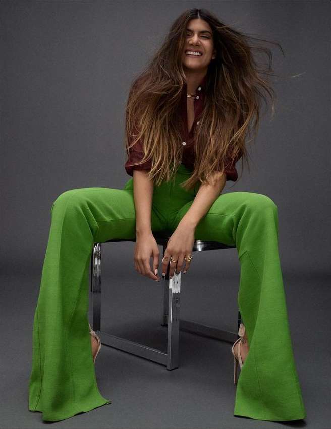 Brunette woman with long hair sits on a metal stool in a photo studio wearing a maroon shirt and bright green bell-bottom pants.