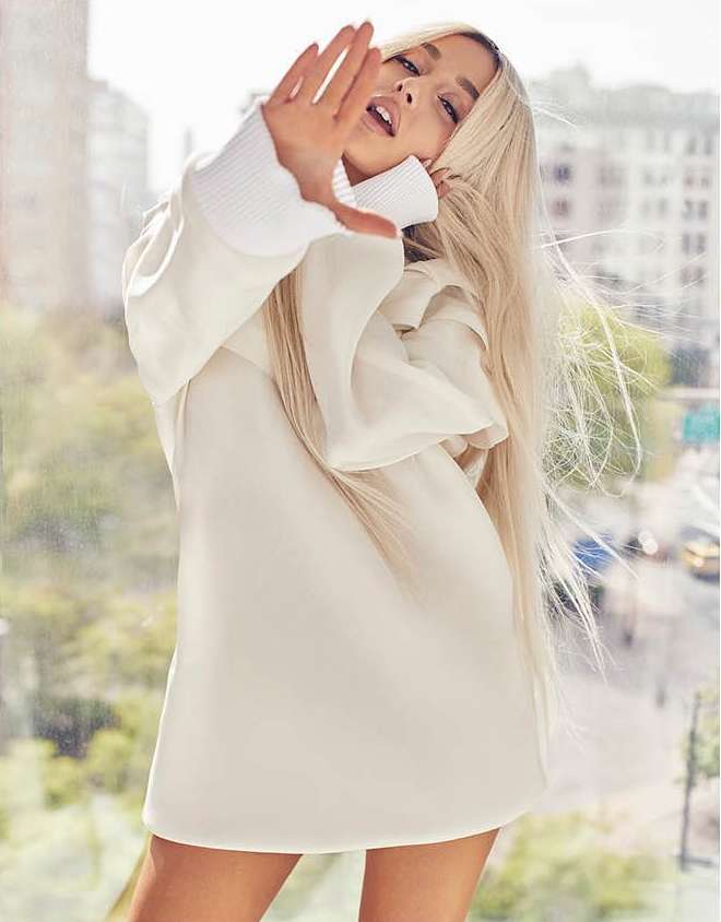Young woman with blonde hair and light skin poses in front of a city backdrop, wearing a short white dress and raising her hand.
