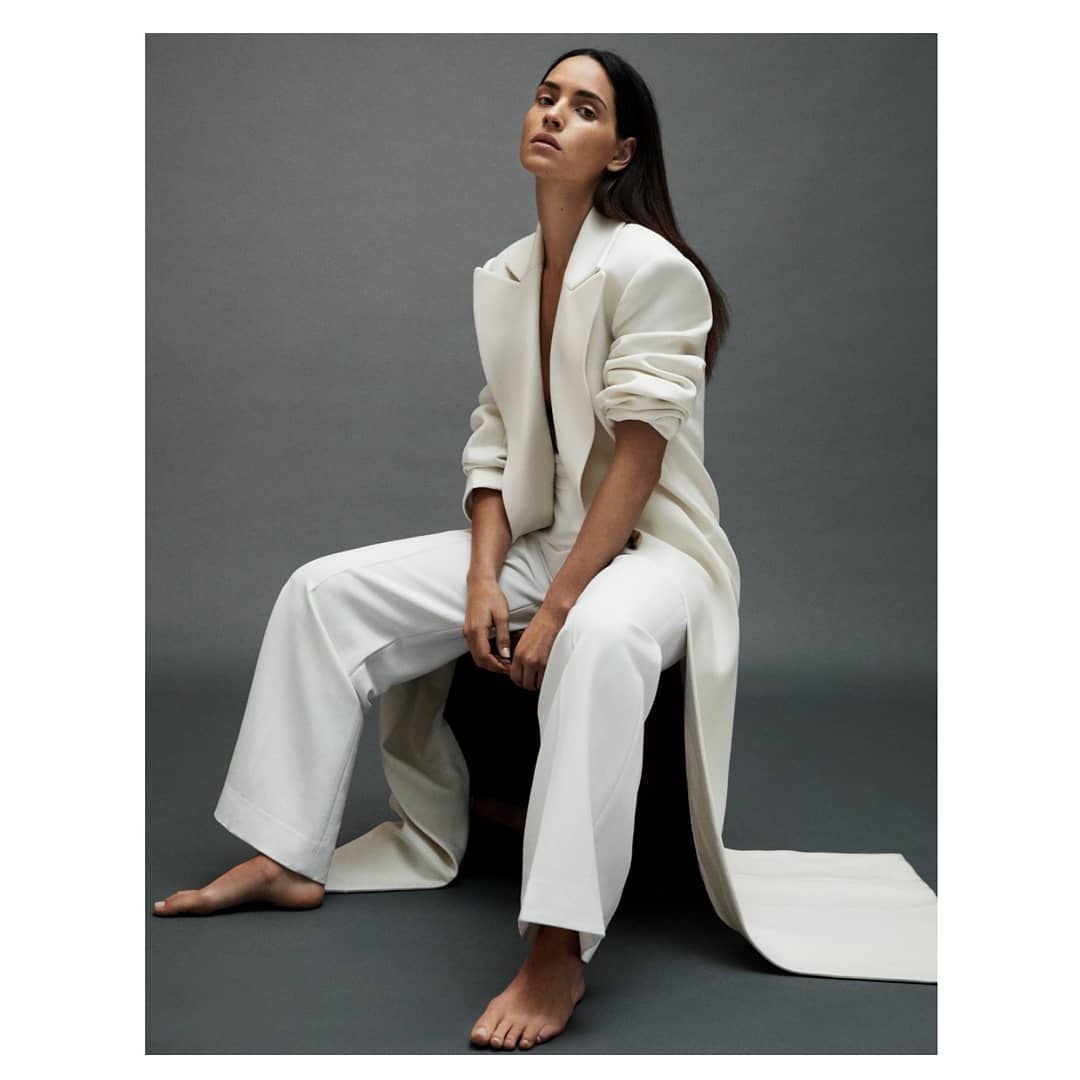 Brunette woman in a white pantsuit poses seated on a chair in a photo studio with neutral grey background. Fashion editorial portrait.