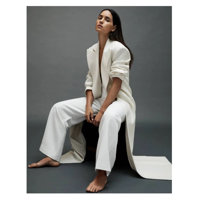 Brunette woman in a white pantsuit poses seated on a chair in a photo studio with neutral grey background. Fashion editorial portrait.