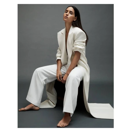 Brunette woman in a white pantsuit poses seated on a chair in a photo studio with neutral grey background. Fashion editorial portrait.