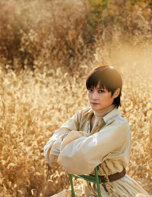 Portrait of a young adult with short dark hair, wearing a tan shirt, sitting on a green chair in a wheat field on a sunny day.