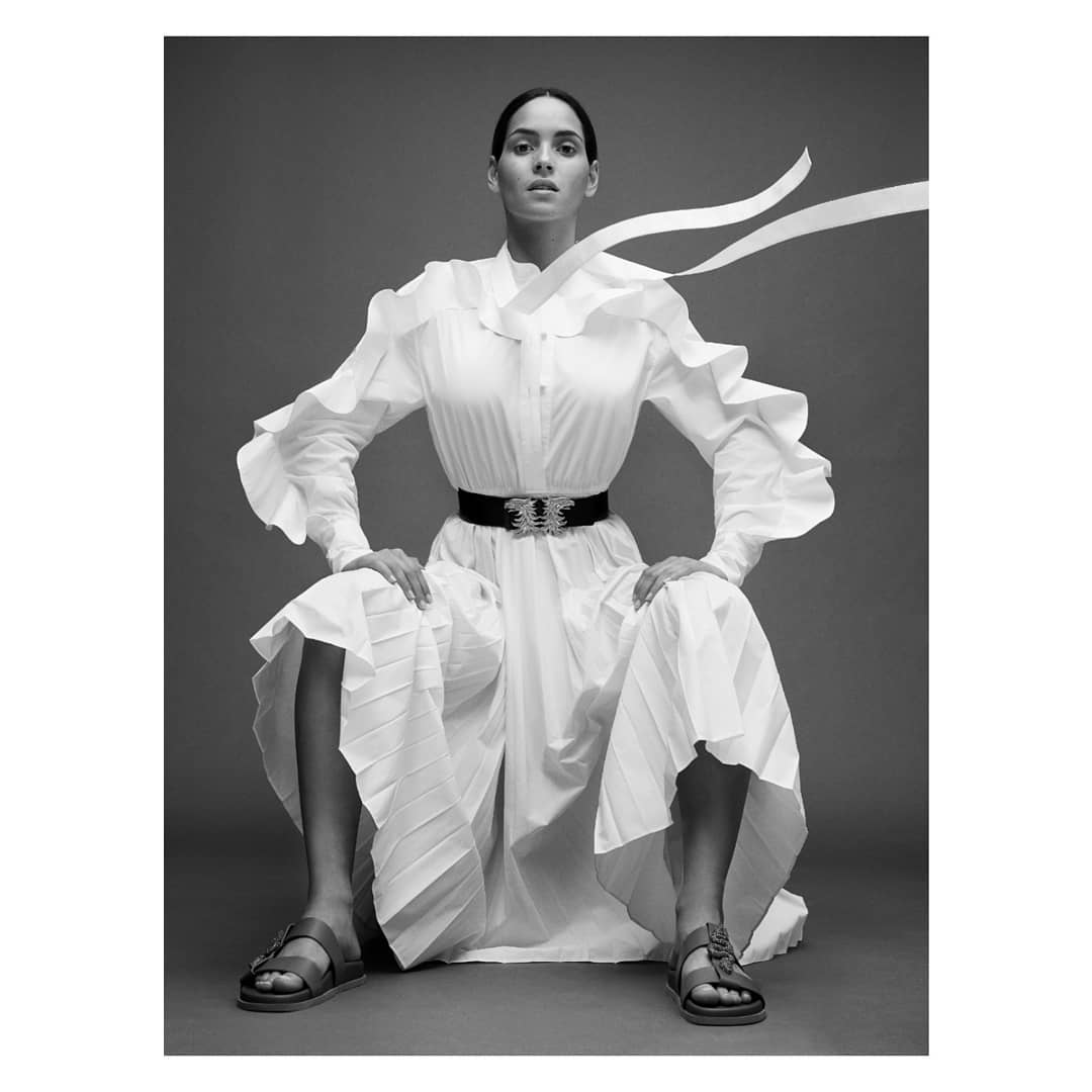 A woman poses in a white dress with a wide belt, seated against a gray backdrop in a studio shot, creating a high fashion look.