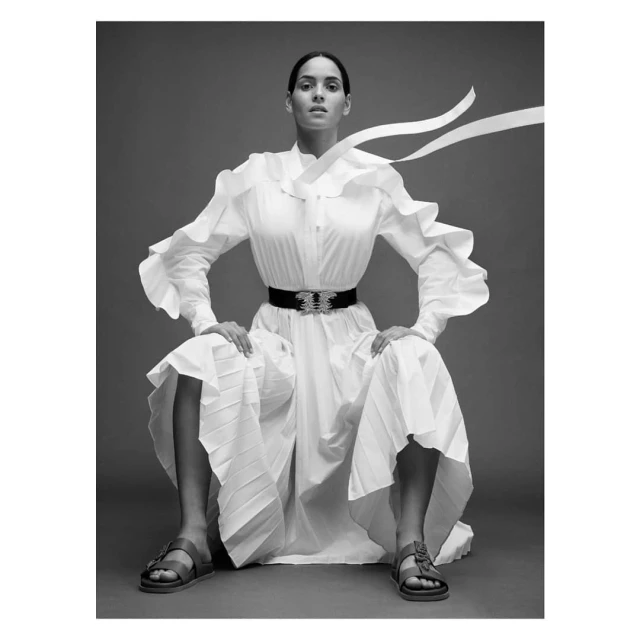A woman poses in a white dress with a wide belt, seated against a gray backdrop in a studio shot, creating a high fashion look.