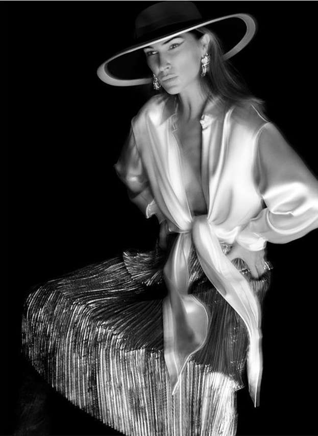 Black and white portrait of a woman in a wide-brimmed hat and satin shirt, seated against a dark backdrop with shiny skirt.