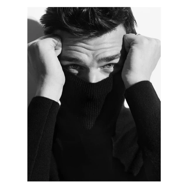 Black and white portrait of an adult male wearing a black turtleneck sweater, with his hands near his face and a bright white background.