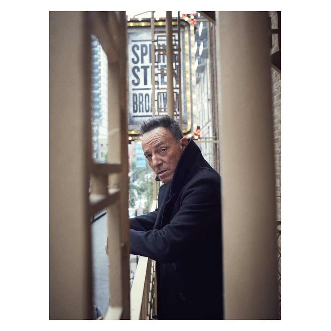 Bruce Springsteen portrait on a balcony, wearing a black coat and looking at the camera with a slight smile and determination.