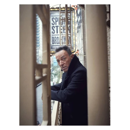 Bruce Springsteen portrait on a balcony, wearing a black coat and looking at the camera with a slight smile and determination.