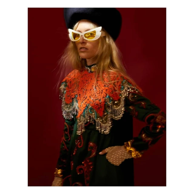 A fair-skinned woman with blonde hair poses in a dark green and orange dress, wearing a black beret and large white sunglasses against a red background.