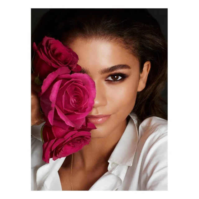 Close-up portrait of a brunette woman with red roses covering part of her face, wearing a white shirt against a dark background.
