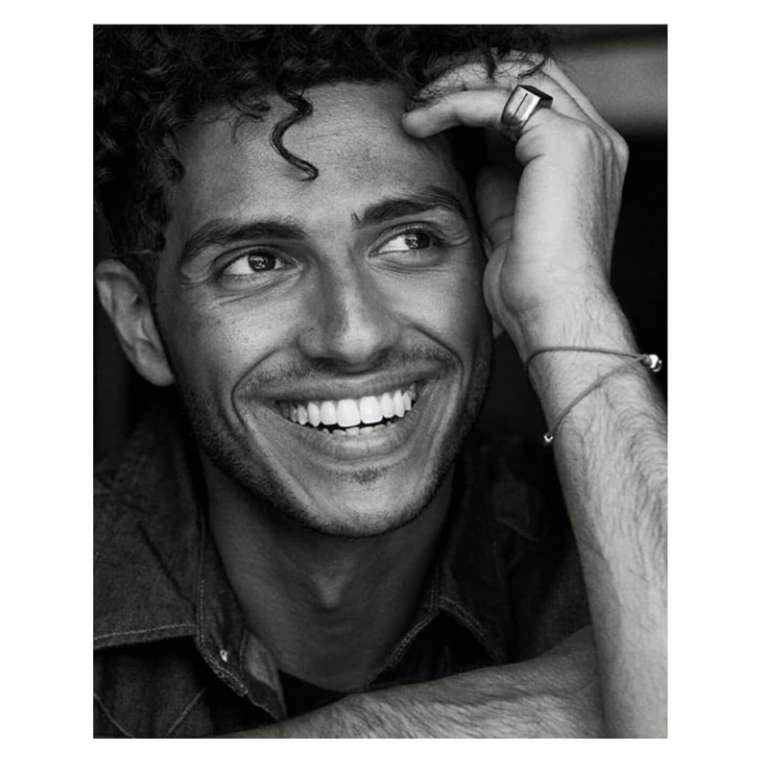Monochromatic portrait of a young adult man with curly hair smiling. He's wearing a denim shirt with a ring and bracelet.