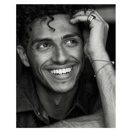 Monochromatic portrait of a young adult man with curly hair smiling. He's wearing a denim shirt with a ring and bracelet.