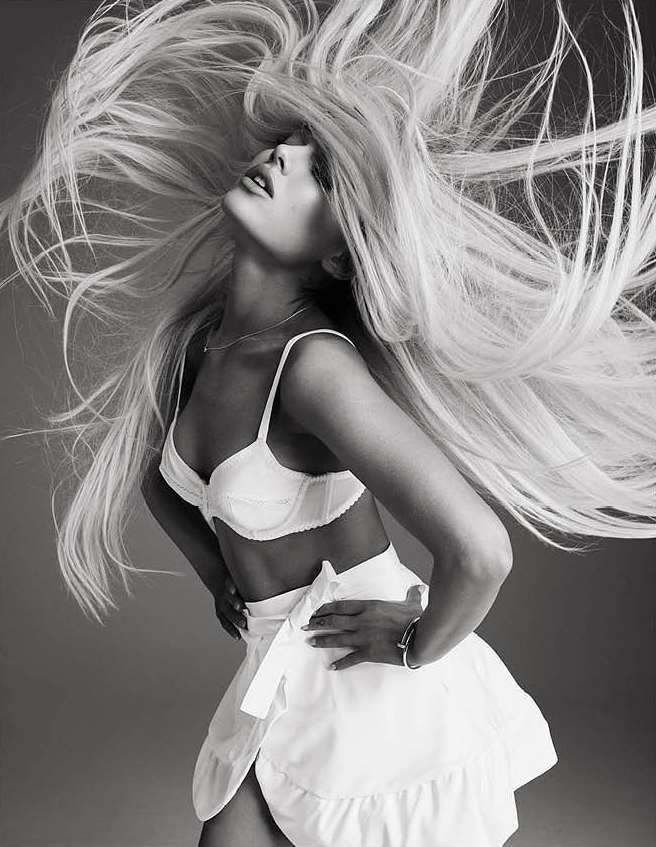 Monochrome portrait of a woman with blonde hair blowing, wearing a white bralette and skirt with a tied bow, against a grey background.