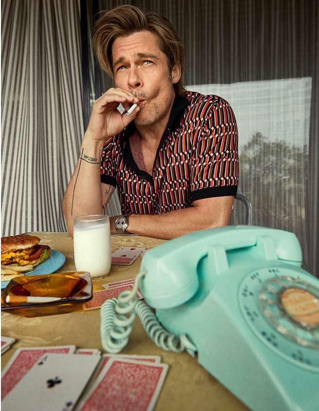 Mature man in patterned shirt smoking indoors at table with playing cards, telephone, milk, and burger; medium shot, shallow depth of field.