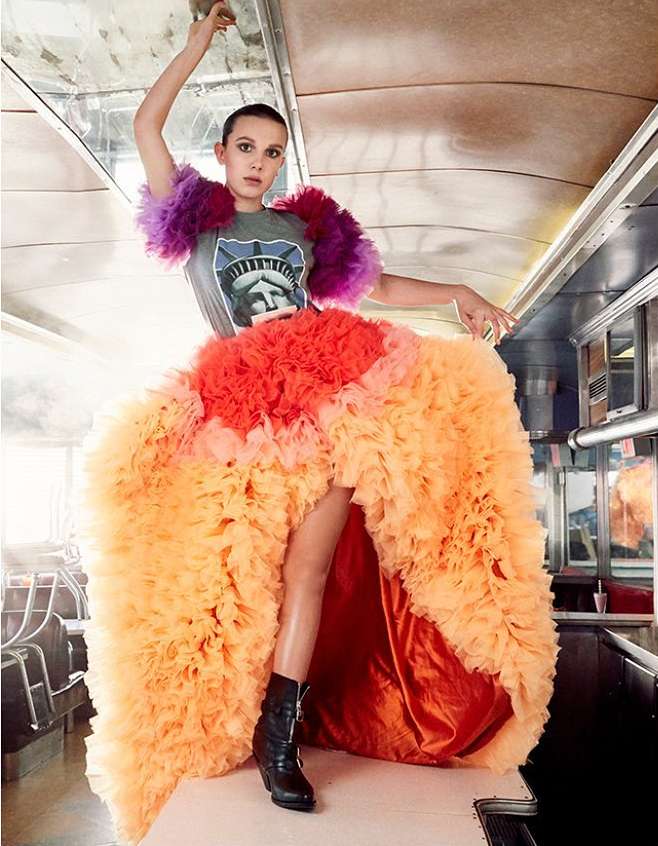 Millie Bobby Brown poses in a sleeveless tee shirt with a Statue of Liberty graphic and a multicolored high-low tulle skirt and black boots.