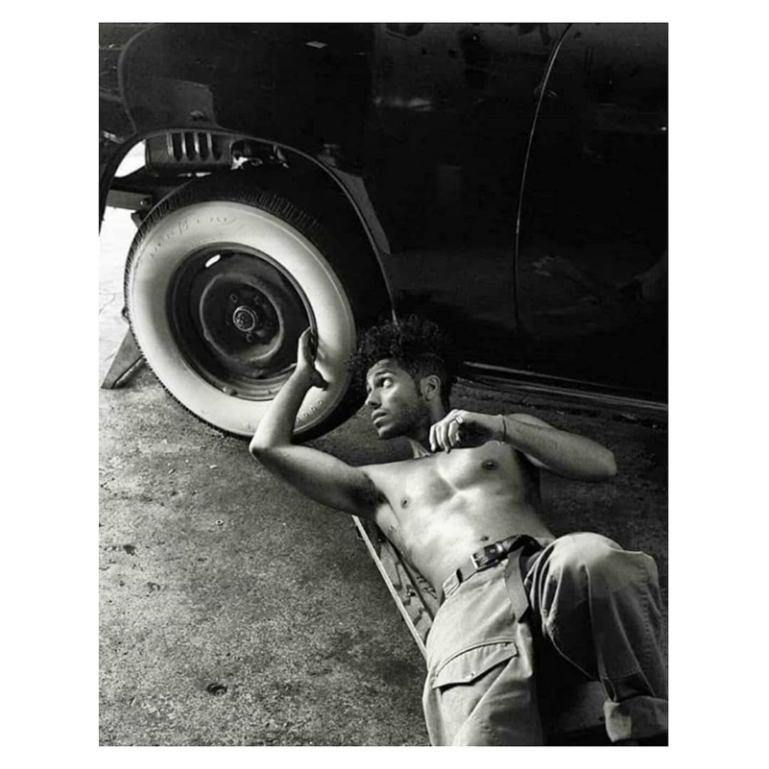 Black and white portrait of a shirtless man with curly hair lying under a vintage car, focusing on the wheel and classic auto design.