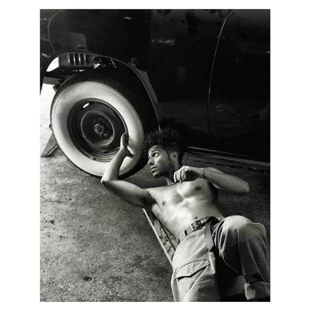 Black and white portrait of a shirtless man with curly hair lying under a vintage car, focusing on the wheel and classic auto design.