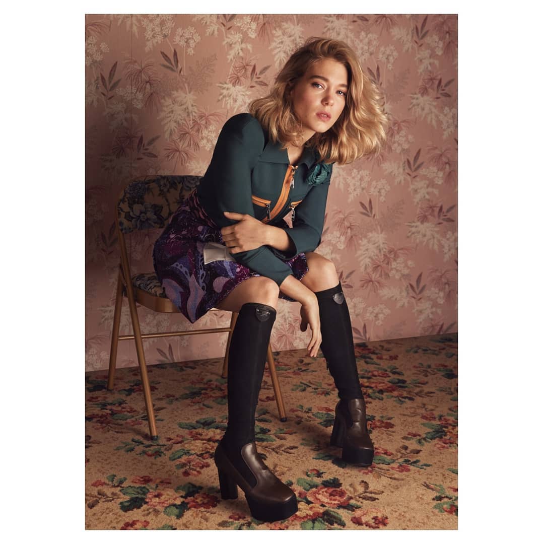 A blonde woman poses seated in tall boots, a short skirt, and green jacket against a floral backdrop, fashion photography.