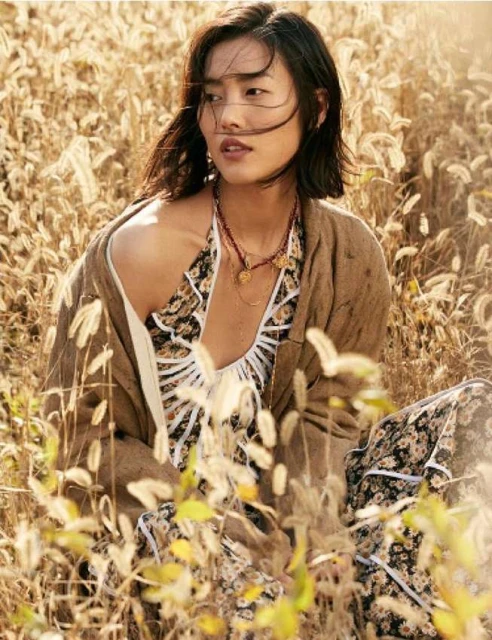Young Asian woman sits in a sunny grassy field wearing a floral print dress and brown cardigan, gazing off to the side in a portrait shot.