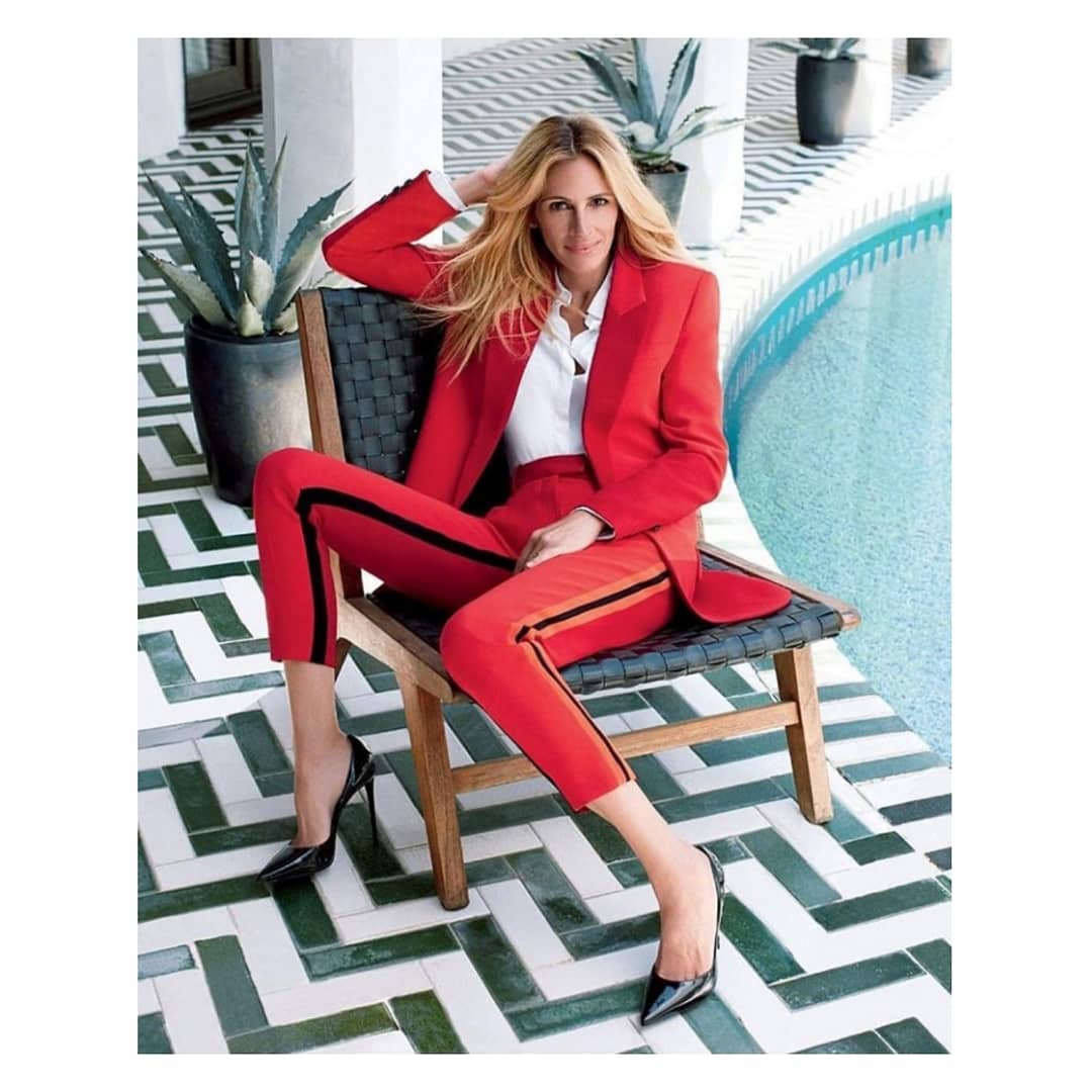 Woman in a red suit sitting on a chair, posing near a swimming pool with a tiled patio and potted plants, creating a fashion editorial image.