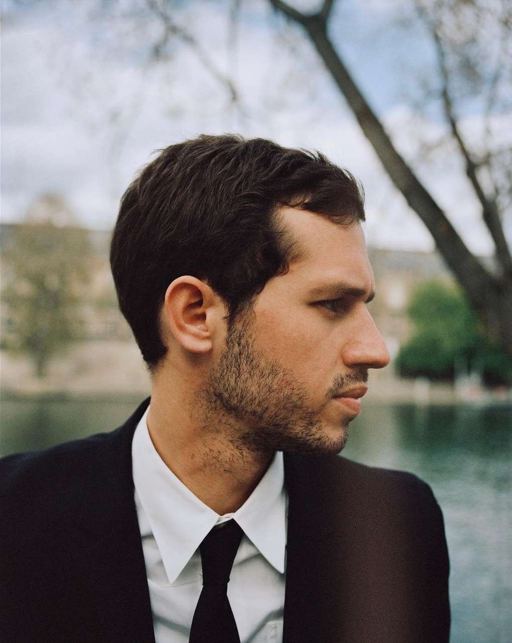 Man in black suit and tie stands outdoors with blurred background. Focus on side profile, showcasing his sharp features and contemplative mood.