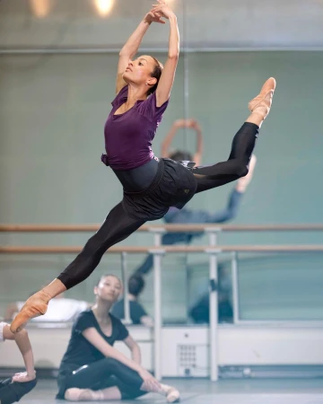 A ballet dancer leaps mid-air in a studio, executing a split with arms raised, showcasing athleticism and grace in a mirrored dance studio setting.