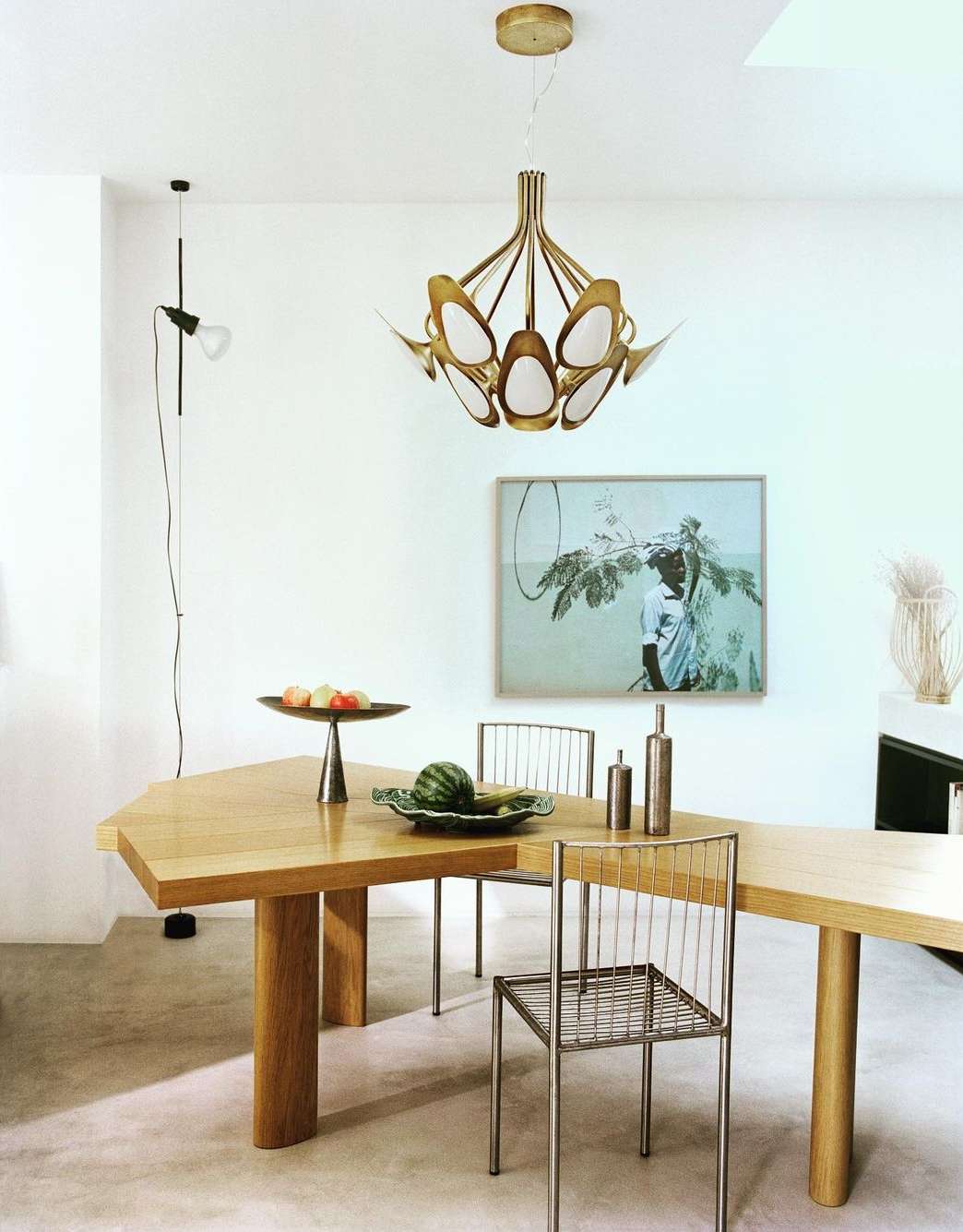 Dining area with wooden table, wire chairs, and unique brass pendant light fixture, set against white walls with framed art.