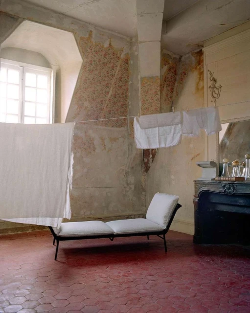 Interior of a rustic room features a chaise lounge, hanging laundry, patterned walls, and red tiled floor, creating a vintage, cozy atmosphere.