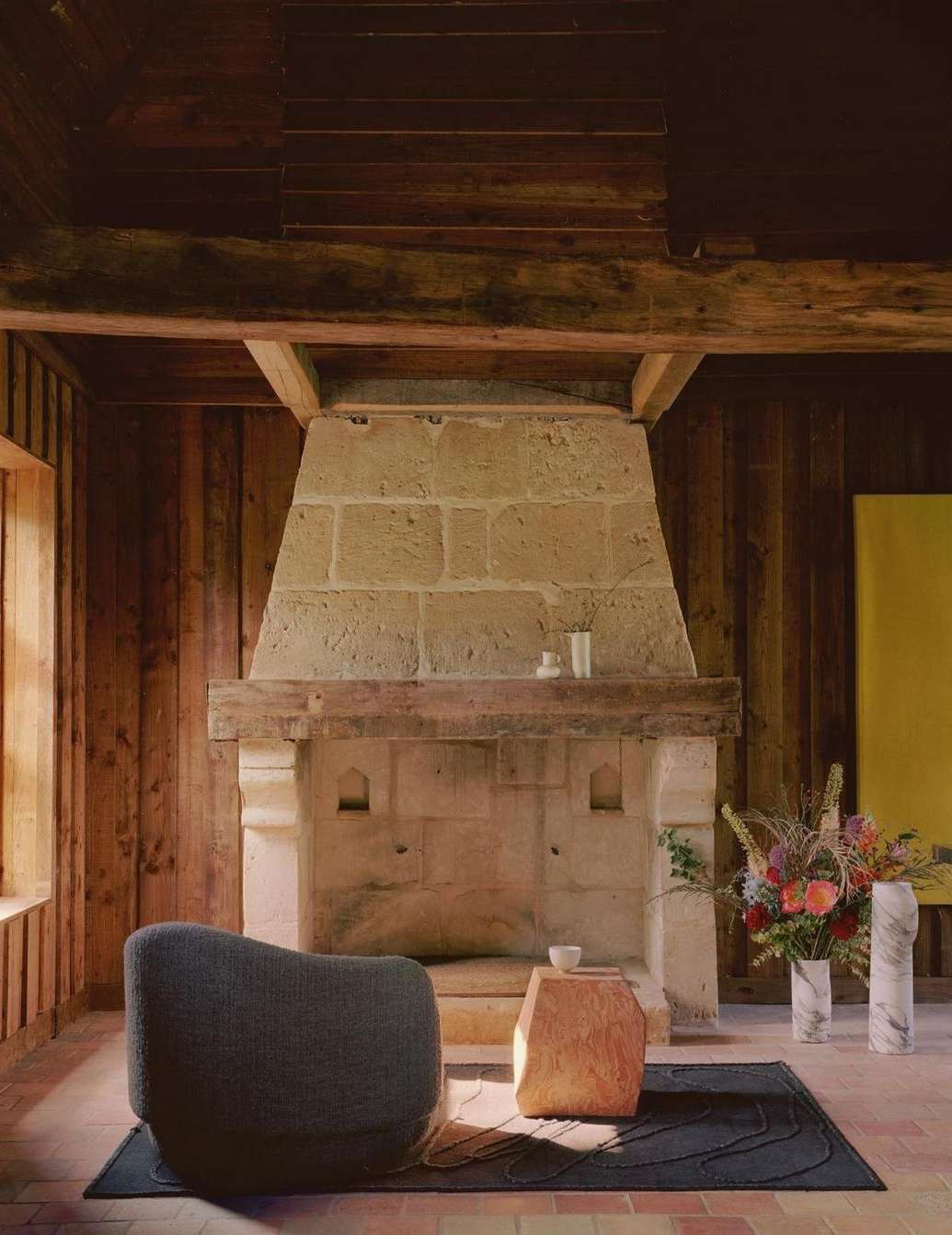 Cozy interior scene with a stone fireplace, modern armchair, sculptural table, and marble vases arranged on a dark-colored rug.